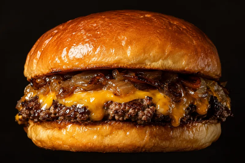 Smashburger med smeltet cheddar og karamelliserede løg i briochebolle, set tæt fra siden mod mørk baggrund