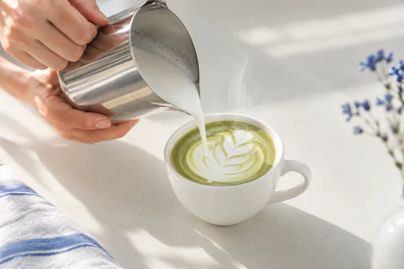 Latte art hældes i en hvid kop på en lys bordplade, set skråt oppefra, damp stiger fra koppen